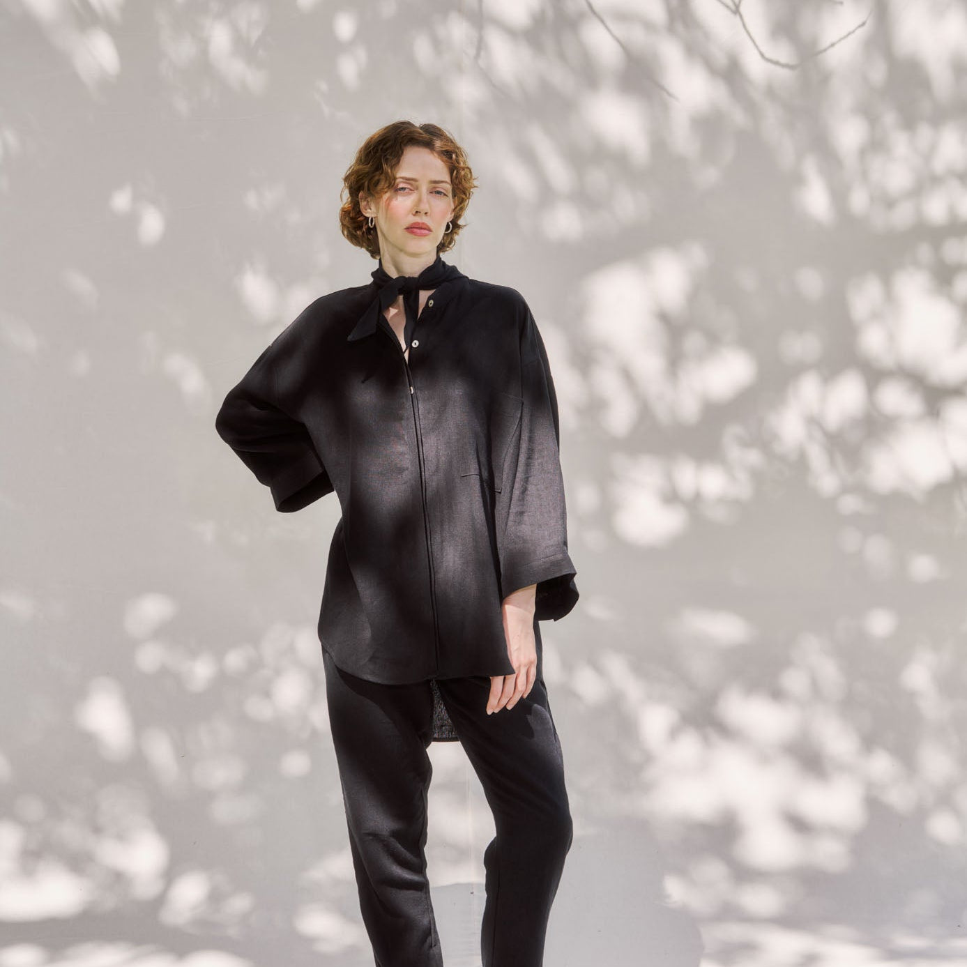 A person standing in front of a textured wall wearing a black blouse with long sleeves and black pants.