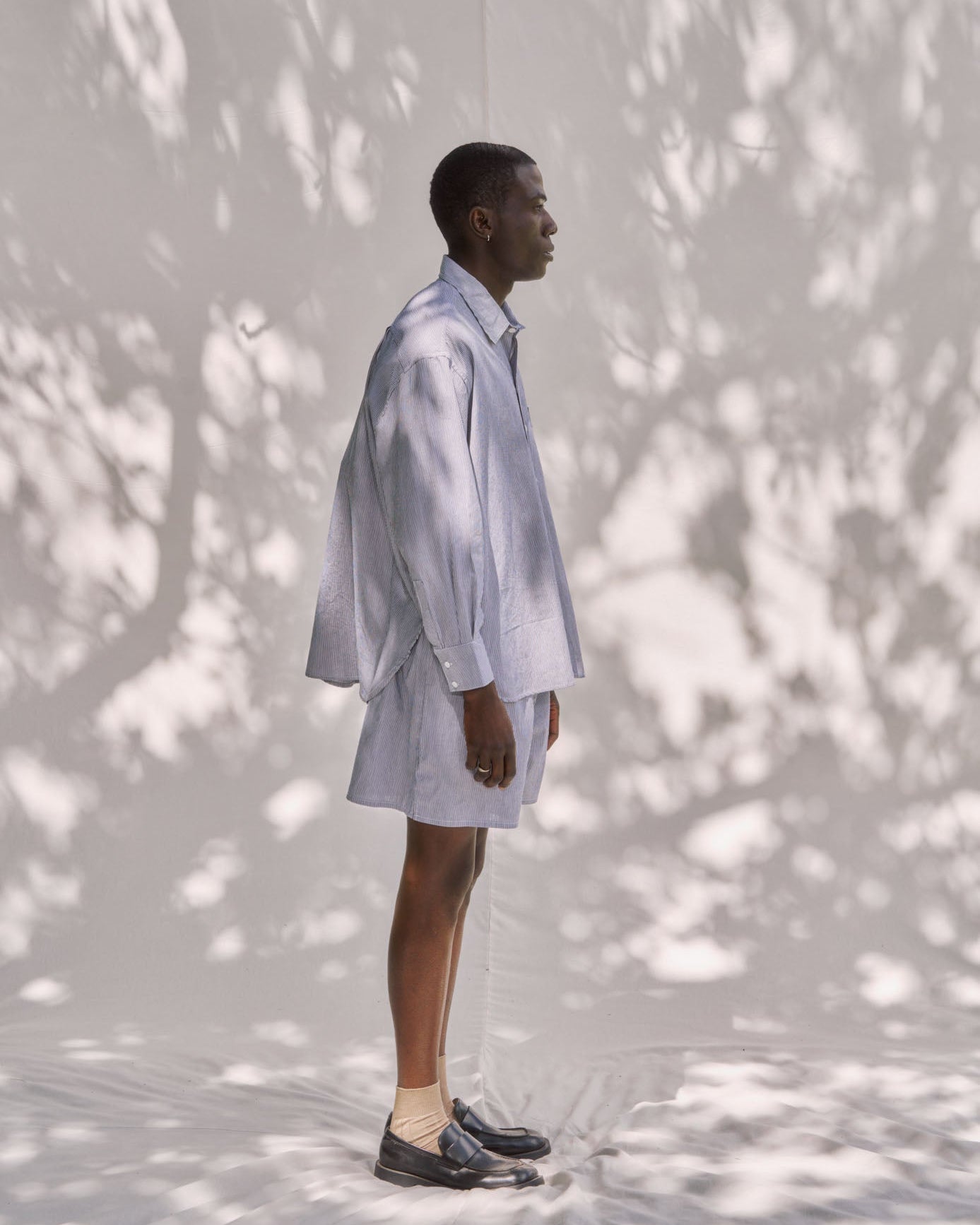 Person wearing a light-colored shirt and pants standing against a white wall with tree shadows.